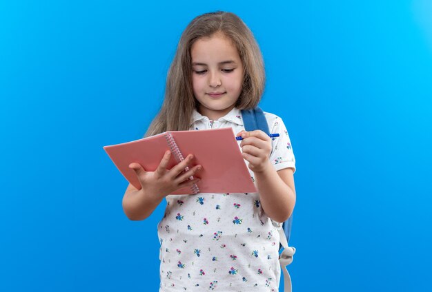 Grave hermosa niña con cabello largo con mochila sosteniendo el cuaderno mirando escribiendo algo con lápiz de pie en azul