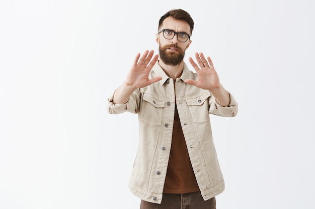 Grave hombre barbudo con gafas posando contra la pared blanca