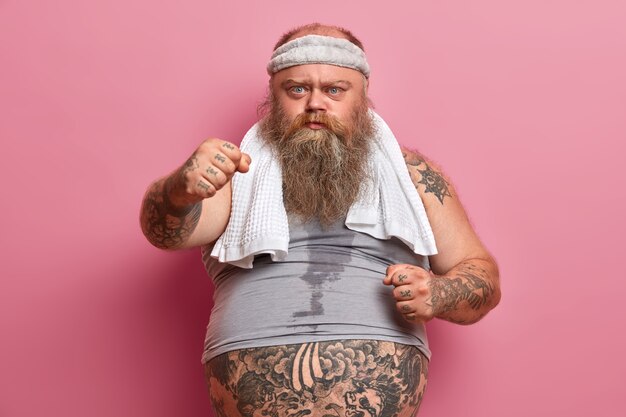 Grave hombre barbudo y regordete muestra los puños cerrados, sufre de sobrepeso, practica deporte, tiene el cuerpo sudoroso y los brazos tatuados, posa contra la pared rosada. Concepto de adelgazamiento y dieta.