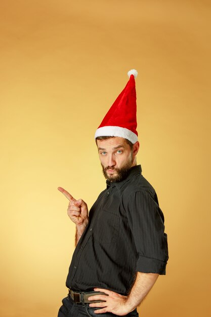 Grave hombre de Navidad con un gorro de Papá Noel en el estudio naranja apuntando a la izquierda