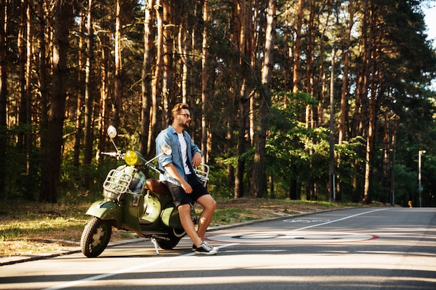 Grave joven barbudo de pie cerca de scooter