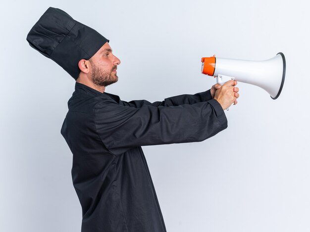 Grave joven cocinero de sexo masculino caucásico en uniforme de chef y gorra de pie en la vista de perfil estirando y mirando al orador aislado en la pared blanca