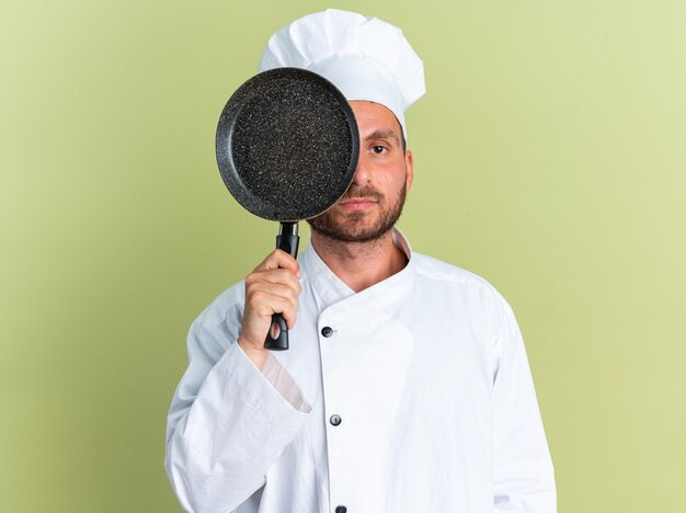 Grave joven cocinero de sexo masculino caucásico en uniforme de chef y gorra que cubre la mitad de la cara con una sartén mirando a la cámara desde atrás aislado en la pared verde oliva