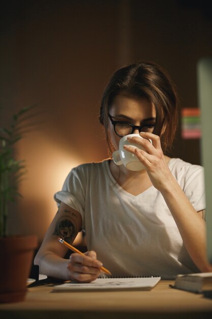 Grave joven diseñadora escribiendo notas y tomando café