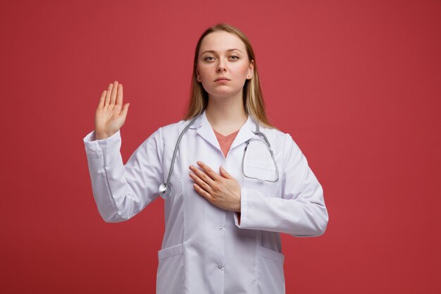 Grave joven doctora rubia vistiendo bata médica y estetoscopio alrededor del cuello haciendo gesto de promesa