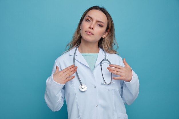 Grave joven doctora vistiendo bata médica y un estetoscopio alrededor del cuello apuntando a sí misma con las manos