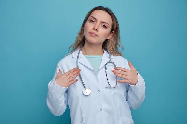 Grave joven doctora vistiendo bata médica y un estetoscopio alrededor del cuello apuntando a sí misma con las manos