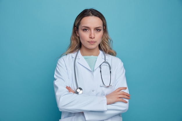 Grave joven doctora vistiendo bata médica y estetoscopio alrededor del cuello de pie con postura cerrada