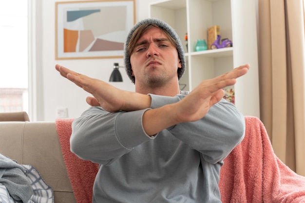 Grave joven enfermo vistiendo gorro de invierno cruzando sus manos gesticulando sin signo sentado en el sofá en la sala de estar