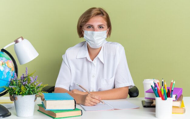 Grave joven estudiante rubia con máscara protectora sentada en el escritorio con herramientas escolares sosteniendo un lápiz mirando a la cámara aislada en la pared verde oliva