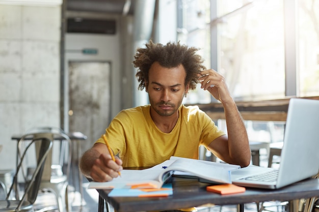 Grave joven estudiante universitario africano enfocado en camiseta amarilla, ocupado haciendo tareas en casa, escribiendo en un libro de ejercicios, sentado en un espacio vacío de coworking temprano en la mañana, usando una computadora portátil