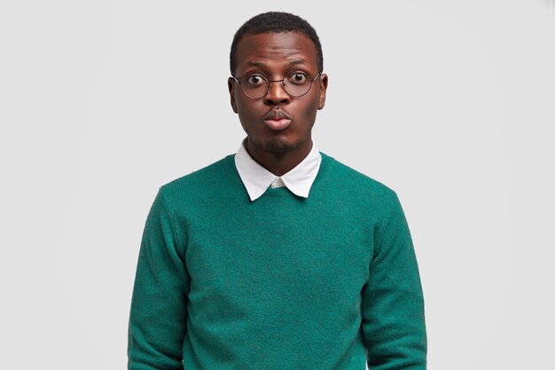 Grave joven hipster masculino redondea los labios para besar, tiene una cita con la mujer, usa gafas redondas, vestido con un suéter verde, aislado sobre fondo blanco.