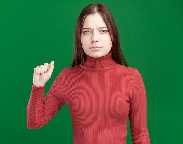 Grave joven mujer bonita mirando al frente haciendo gesto de golpe aislado en la pared verde