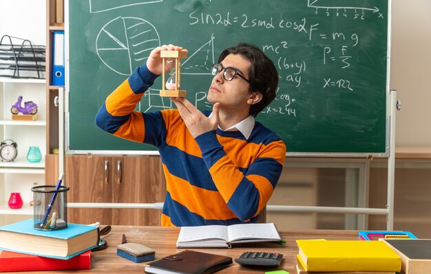 Grave joven profesor de geometría caucásico con gafas sentado en un escritorio con herramientas escolares en el aula sosteniendo el reloj de arena mirándolo verticalmente