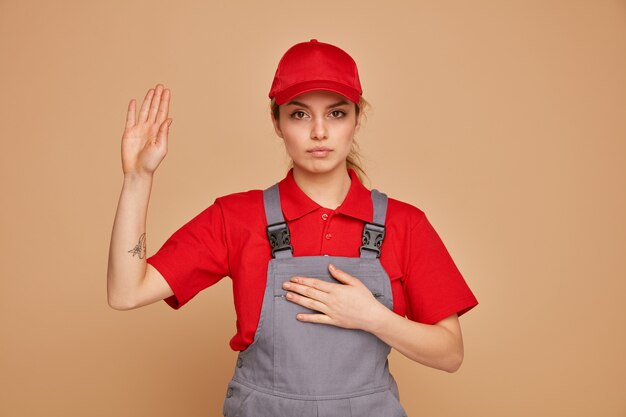 Grave joven trabajador de la construcción con uniforme y gorra haciendo gesto de promesa