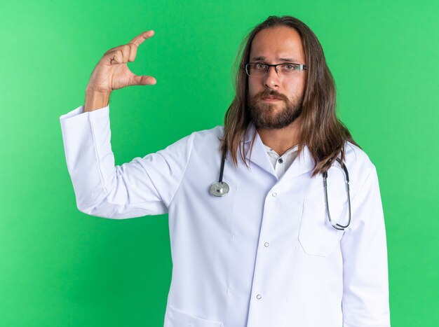 Grave médico varón adulto vistiendo bata médica y un estetoscopio con gafas mirando a la cámara haciendo un gesto de pequeña cantidad aislado en la pared verde