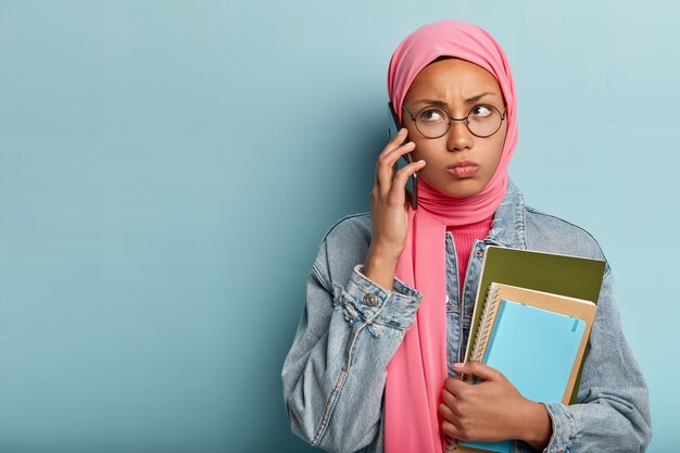 Grave mujer árabe llama por teléfono celular, enfocada a un lado, tiene expresión facial gruñona, usa anteojos redondos