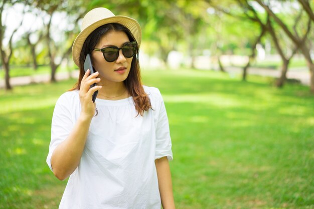 Grave mujer asiática joven pensativa en gafas de sol hablando por teléfono móvil