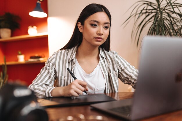 Grave mujer asiática de ojos marrones que trabaja en la oficina con un portátil y sosteniendo la pluma