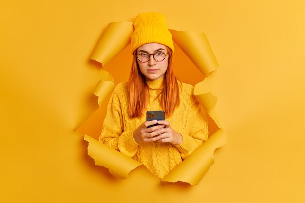 Grave mujer bonita pelirroja con smartphone, viste suéter y sombrero amarillo, rompe la pared de papel