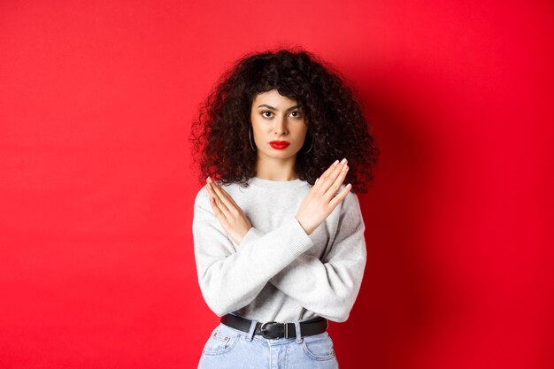 Grave mujer caucásica con cabello rizado dice que no, haciendo un gesto cruzado para detener o prohibir algo malo, de pie disgustado sobre fondo rojo.