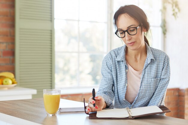 Grave mujer caucásica lleva gafas y camisa, menú creats como se sienta sobre el interior de la cocina.