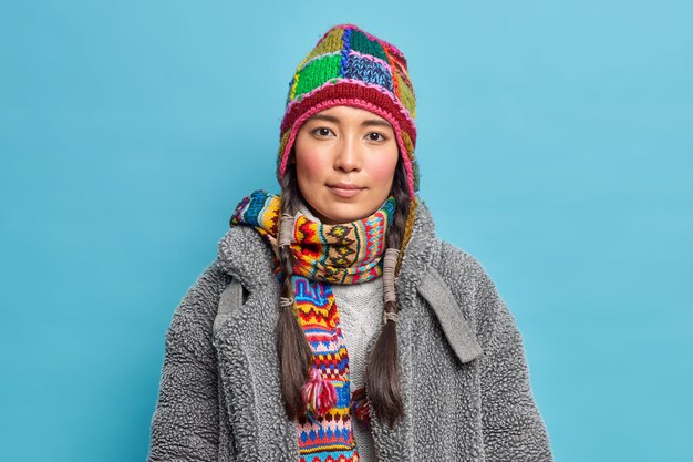 Foto gratuita grave mujer escandinava con coletas mira tranquilamente al frente vestida con ropa de invierno abrigada posa sobre la pared azul