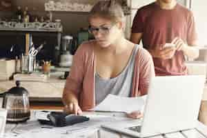 Foto gratuita grave mujer joven con gafas rectangulares calculando gastos mientras realiza el presupuesto familiar usando una computadora portátil genérica y una calculadora en casa