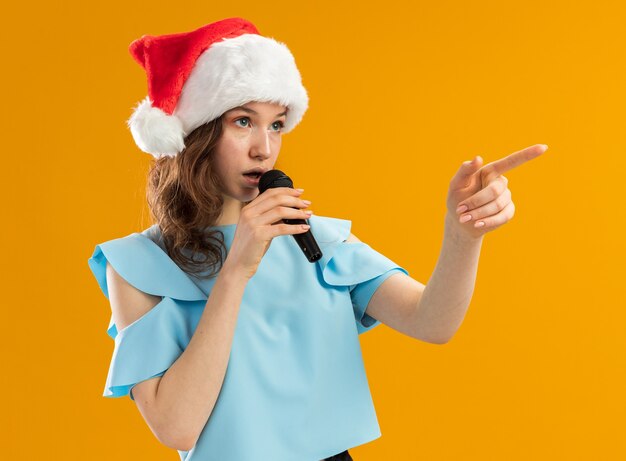 Grave mujer joven en la parte superior azul y gorro de Papá Noel hablando con el micrófono apuntando con el dedo índice hacia el lado