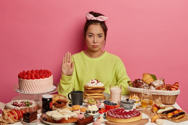 Grave mujer morena hace gesto de rechazo, posa junto a la mesa de la cocina con postres, sigue la dieta, se niega a comer alimentos con muchas calorías. Dulces de moda