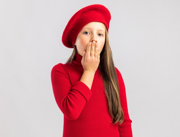 Grave niña rubia con boina roja mostrando gesto de sorpresa aislado en la pared blanca con espacio de copia
