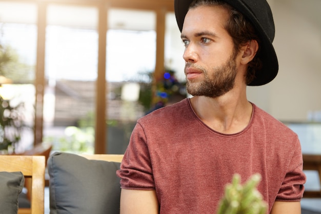 Grave o triste atractivo joven estudiante barbudo con sombrero negro de moda sentado solo en la moderna cafetería espaciosa