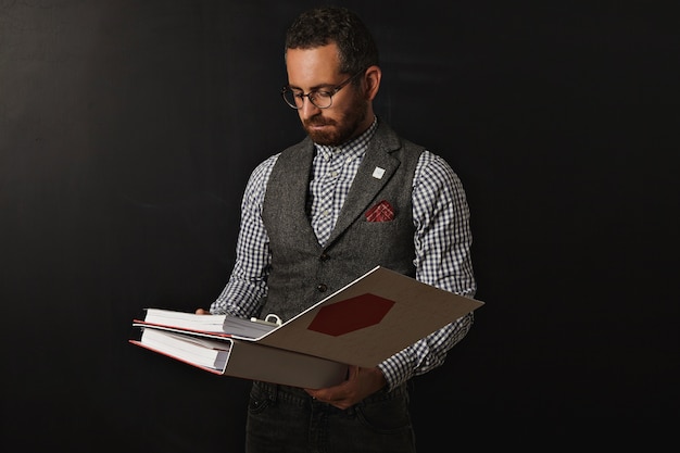 Grave profesor barbudo con camisa oxford a cuadros y chaleco de tweed, con gafas lee el plan educativo en dos grandes carpetas de documentos para su estudiante para el próximo año en la universidad