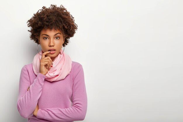 Grave señorita rizada con cabello afro oscuro, mantiene los brazos parcialmente cruzados, viste poloneck morado