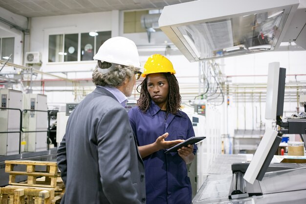 Grave trabajadora de fábrica hablando con el jefe en la máquina en el piso de la planta, apuntando a la pantalla de la tableta