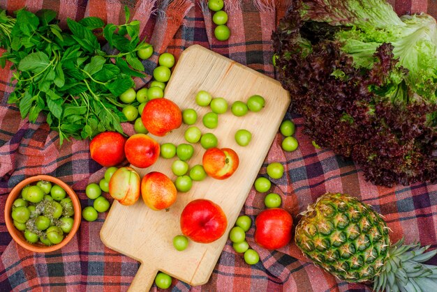 Greengages y duraznos dispersos con hojas verdes, una piña y lechuga en una tabla de cortar y un tazón de arcilla sobre tela de picnic, en posición plana.