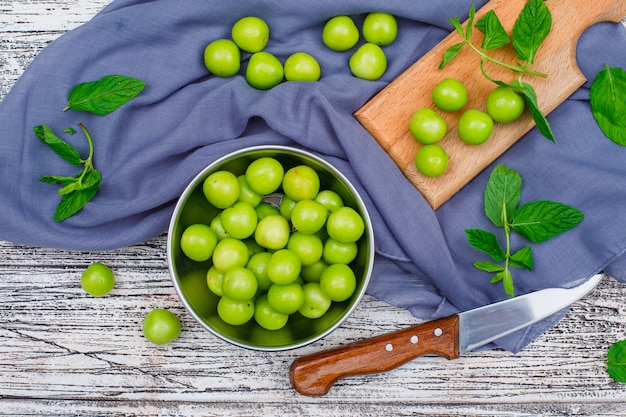 Greengages con hojas en una cacerola de metal y tabla de cortar de madera con cuchillo plano sobre madera gris y tela de picnic