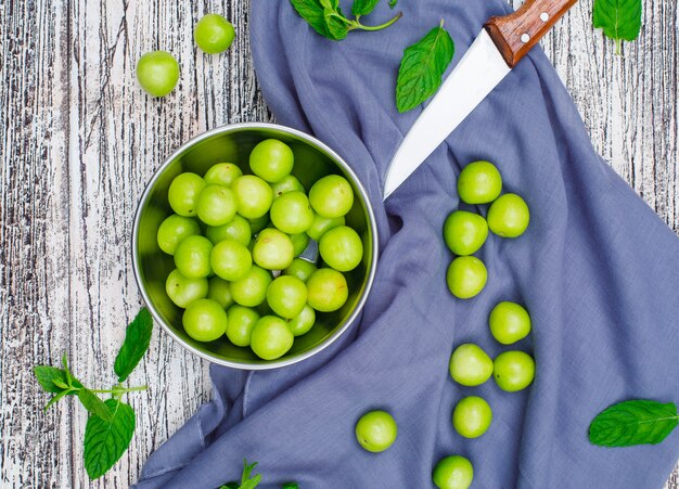 Greengages con hojas con cuchillo en una cacerola de metal sobre madera gris y tela de picnic, vista de ángulo alto.