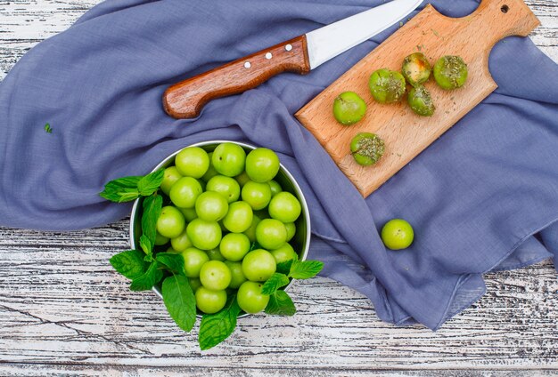 Greengages picantes con hojas en una cacerola de metal y tabla de cortar de madera con cuchillo vista de ángulo alto sobre una madera gris y tela de picnic