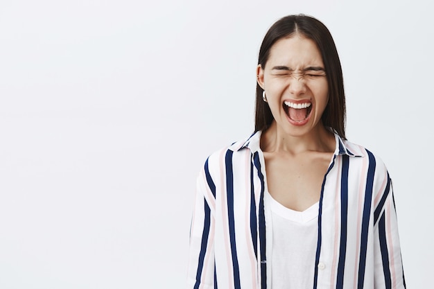 Grita como nadie oye. Retrato de mujer emprendedora elegante y atractiva harta en blusa a rayas, cerrando los ojos y gritando, deprimida y aliviada del estrés, gritando en voz alta