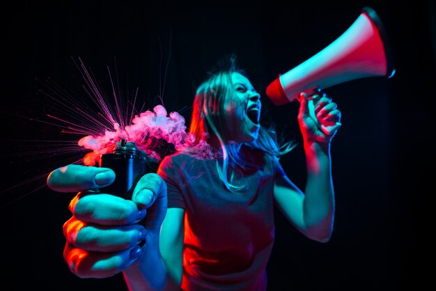 Gritando con megáfono. Mujer joven con humo y luz de neón sobre fondo negro. Vista de ojo de pez muy tensada, gran angular.