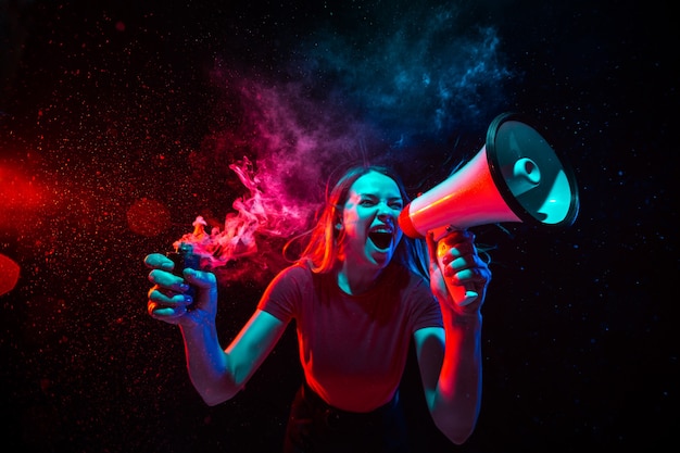 Gritando con megáfono. Mujer joven con humo y luz de neón sobre fondo negro. Vista de ojo de pez muy tensada, gran angular.