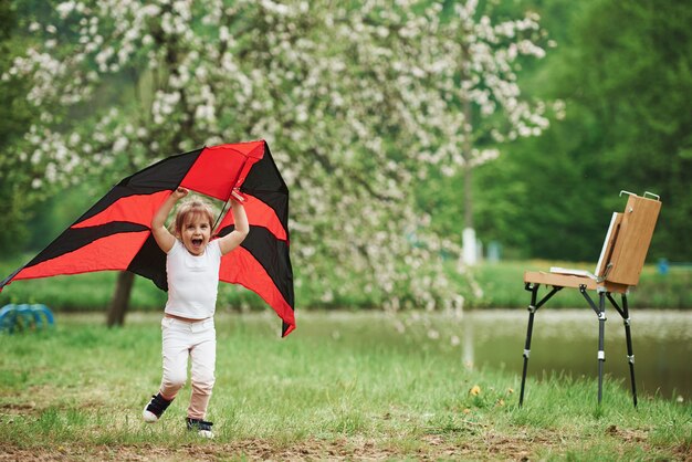 Grito de felicidad. Niña positiva corriendo con cometa de color rojo y negro en las manos al aire libre