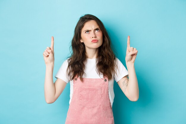 Gruñón linda chica morena frunciendo el ceño disgustado, enfurruñado y mirando decepcionado, señalando con el dedo en la parte superior, de pie contra el fondo azul.