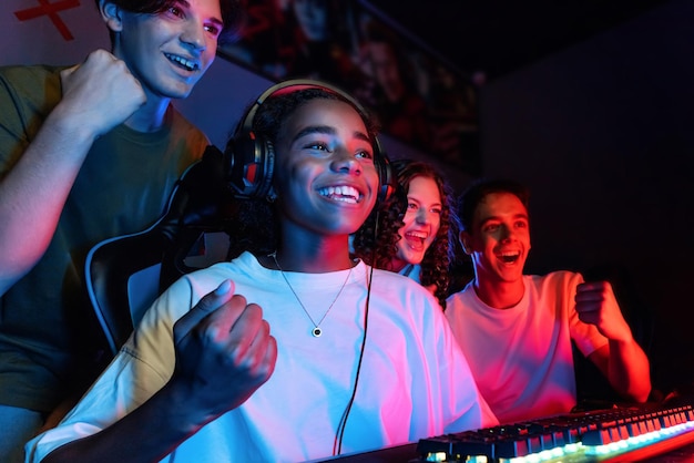 Grupo de adolescentes multirraciales jugando videojuegos en un club de videojuegos con iluminación azul y roja emocionados por una victoria