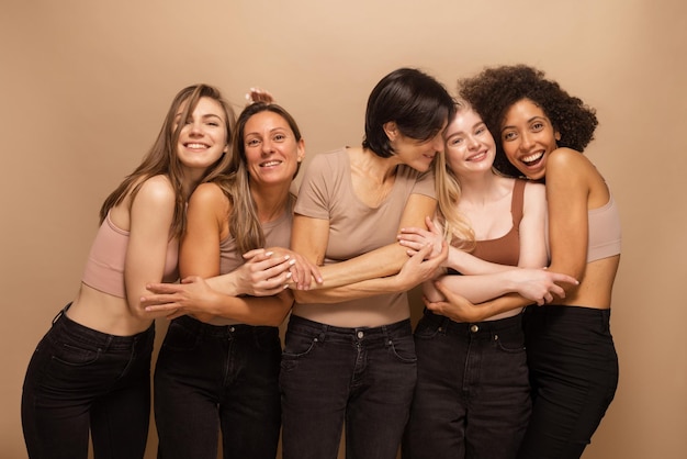 Grupo de alegres mujeres interraciales con blusas marrones y pantalones negros de pie juntas frente a un fondo marrón Novias multiétnicas en el estudio