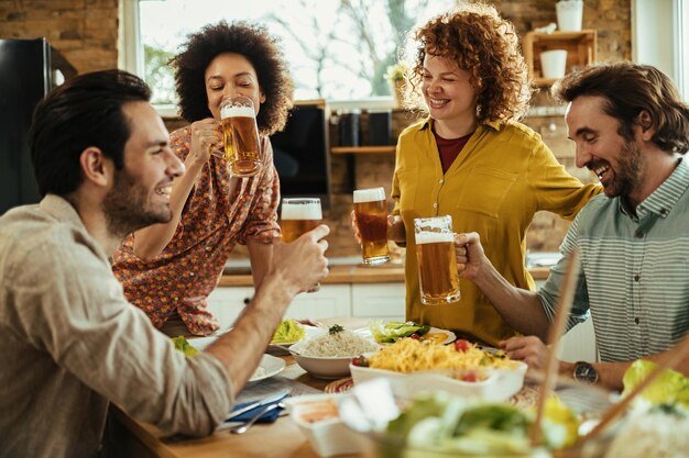 Grupo de amigos alegres bebiendo cerveza y divirtiéndose mientras comen en la mesa del comedor.