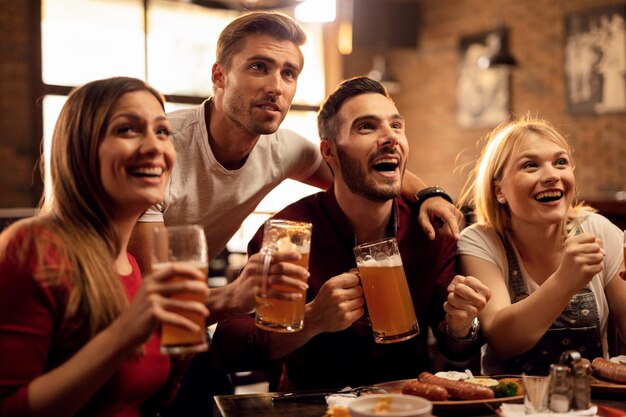 Grupo de amigos alegres divirtiéndose mientras ven juegos deportivos en la televisión y beben cerveza en un bar