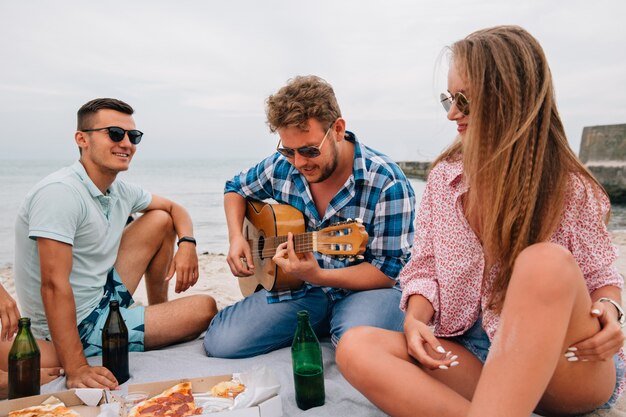 Grupo de amigos atractivos haciendo un picnic, tocando la guitarra en la playa, mientras come pizza