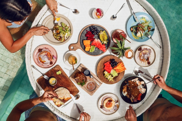 Grupo de amigos desayunando tropical en bandeja flotante en la piscina. Frutas y bebidas exóticas frescas. Ambiente de fiesta.